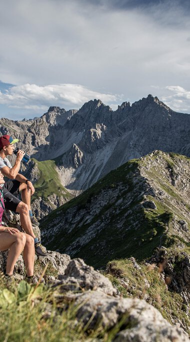 Gruppe von Wanderern auf einem bergigen Gipfel mit Blick auf beeindruckende Bergketten. | © Kleinwalsertal Tourismus eGen • Fotograf: Bastian Morell