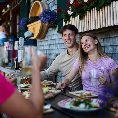 Fröhliche Menschen beim Essen im Außenbereich einer Alpe, umgeben von Blumen und Holzwänden, im Sommer 2024. | © Kleinwalsertal Tourismus eGen • Fotograf: Oliver Farys