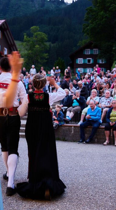 Traditionelle Trachtengruppe beim Sommerfestival in Kleinwalsertal, tanzend vor großem Publikum im Freien bei Dämmerung. | © Kleinwalsertal Tourismus eGen • Fotograf: Oliver Farys
