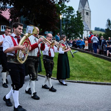 Blasmusikkapelle in traditioneller Tracht beim Umzug in Mittelberg während des Sommerfestivals, mit Publikum im Hintergrund und einem historischen Kirchturm. | © Kleinwalsertal Tourismus eGen • Fotograf: Oliver Farys