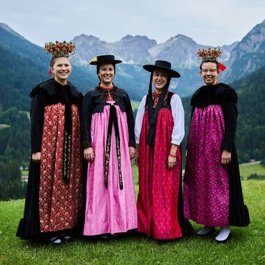 Vier Frauen in traditioneller Tracht stehen auf einer grünen Wiese vor einer bergigen Landschaft mit Wäldern und Bergen im Hintergrund. | © Kleinwalsertal Tourismus eGen • Fotograf: Oliver Farys