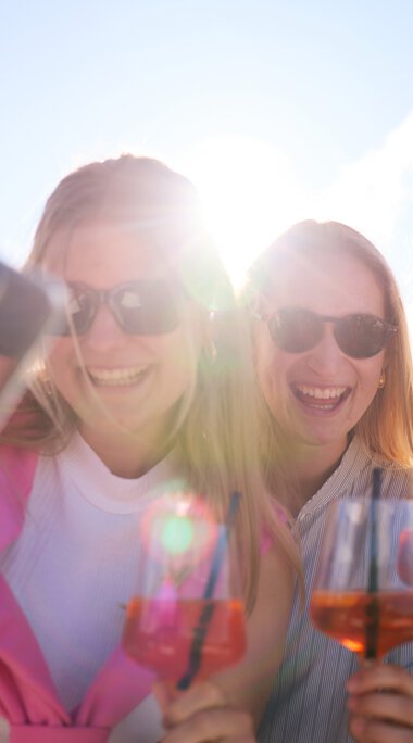 Drei Frauen machen ein Selfie im Freien bei sonnigem Wetter, lachen und haben Getränke in der Hand. | © Kleinwalsertal Tourismus eGen • Fotograf: Oliver Farys