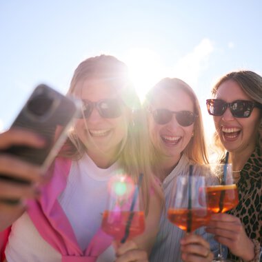 Drei Frauen machen ein Selfie im Freien bei sonnigem Wetter, lachen und haben Getränke in der Hand. | © Kleinwalsertal Tourismus eGen • Fotograf: Oliver Farys