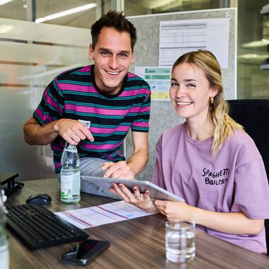 Zwei junge Menschen in einem modernen Büro, lachend während einer Arbeitssitzung, mit einem Computer und Dokumenten auf dem Tisch. | © Kleinwalsertal Tourismus eGen • Fotograf: Oliver Farys