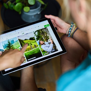 Zwei Personen betrachten Fotos auf einem Tablet, die draußen aufgenommen wurden, mit Fokus auf eine Damengeburtstagsfeier im Grünen. | © Kleinwalsertal Tourismus eGen • Fotograf: Oliver Farys