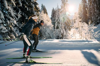 Zwei Skifahrer beim Langlaufen durch eine verschneite Winterlandschaft im Kleinwalsertal, Sonnenlicht durch Bäume, klare Winterluft. | © Kleinwalsertal Tourismus eGen • Fotograf: Oliver Farys