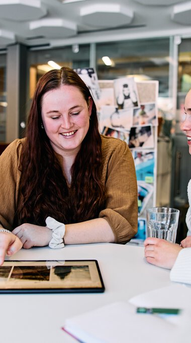 Vier junge Frauen sitzen um einen Tisch, lachen und schauen auf ein Tablet, in einem modernen Büro. | © Kleinwalsertal Tourismus eGen • Fotograf: Oliver Farys