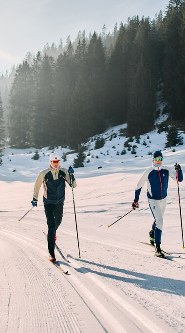 Zwei Skilangläufer in winterlicher Landschaft beim Langlaufen auf der Schwarzwasserloipe im Kleinwalsertal, umgeben von Tannen und verschneiten Bergen. | ©  Kleinwalsertal Tourismus eGen • Fotograf: Oliver Farys