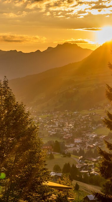 Bergsommer bei Sonnenuntergang mit Blick auf die Berge, Täler und eine kleine Siedlung, umgeben von Bäumen im goldenen Licht. | © Kleinwalsertal Tourismus eGen • Fotograf: Steffen Berschin 