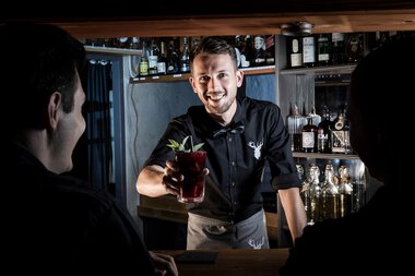 Barista oder Barkeeper serviert einen roten Cocktail an zwei Gäste in einer Bar mit Getränkeflaschen im Hintergrund. | © Kleinwalsertal Tourismus eGen • Fotograf: Werner Krug