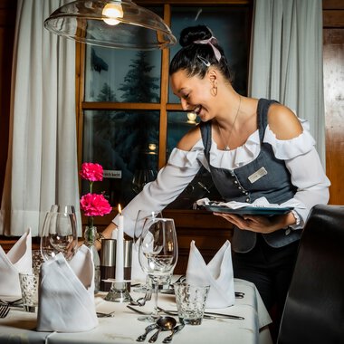 Eine freundliche oder professionelle Kellnerin bereitet einen Tisch in einem Restaurant vor, mit schön gedeckten Tischen, Kerzen und Gläsern. | © Kleinwalsertal Tourismus eGen • Fotograf: Werner Krug
