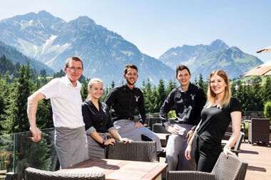 Fünf Mitarbeiter des Walserhofs sitzen auf einer Terrasse vor einer Bergkulisse mit Bergen und Wäldern, im sonnigen Wetter. | © Kleinwalsertal Tourismus eGen • Fotograf: Werner Krug