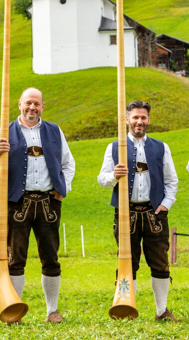 Vier Männer in traditioneller Tracht stehen im Grünen, jeder hält ein Alphorn, bei einem Alphornfestival im Jahr 2022. | © Kleinwalsertal Tourismus eGen • Fotograf: Frank Drechsel
