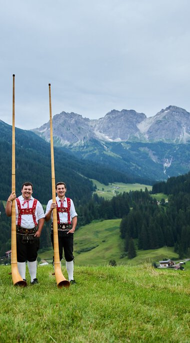 Drei Männer in Trachten stehen in einer grünen Wiese mit Alphörnern, umgeben von Bergen und einer bewaldeten Talregion. | © Kleinwalsertal Tourismus eGen • Fotograf: Oliver Farys